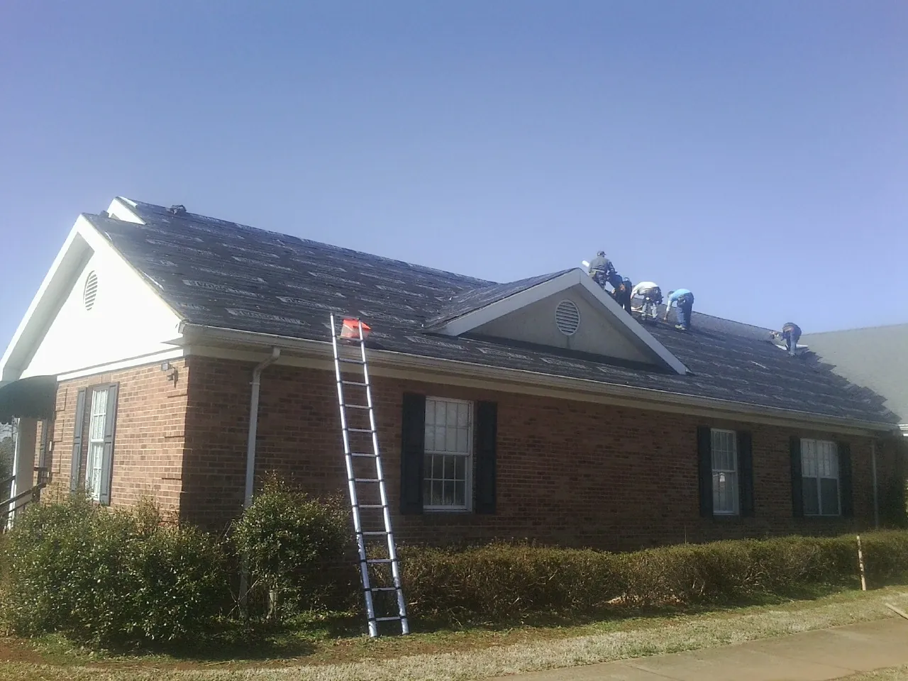 Crew tearing shingles off a brick ranch roof with a ladder set — Emergency Roof Repair work by our crew in Imler
