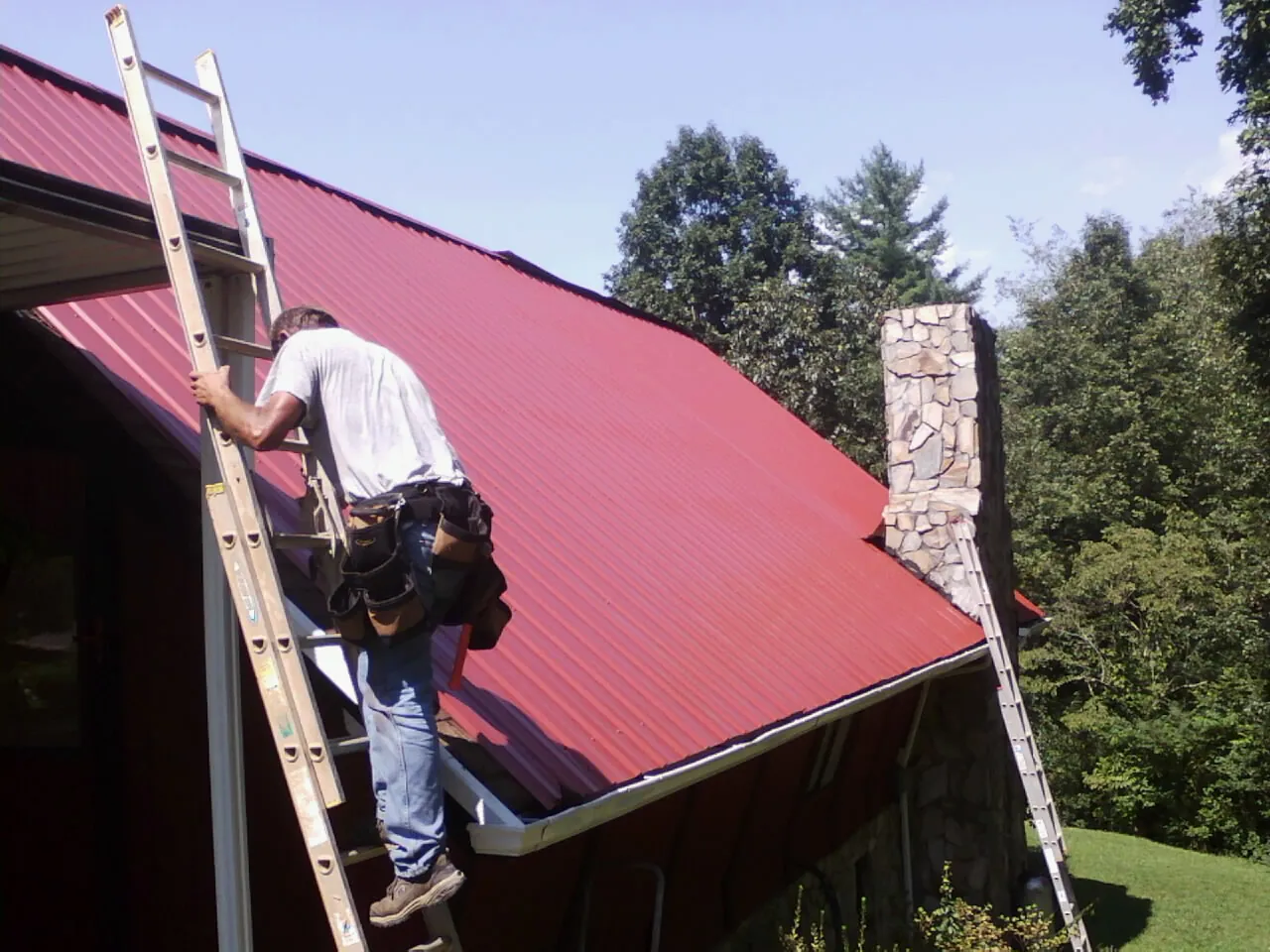 Roofer on a ladder beside a red metal roof and stone chimney — Soffit & Fascia Repair by Roofing Brothers in Imler